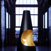 Archimedes Kamin – hoher schwarzer Standkamin mit goldenem Innenleben und Flamme in einer Halle